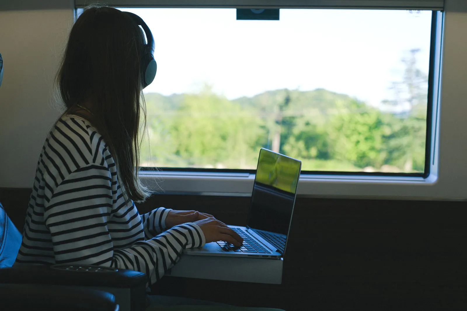 Woman using a laptop with headphones during a train journey, enjoying the scenic view.